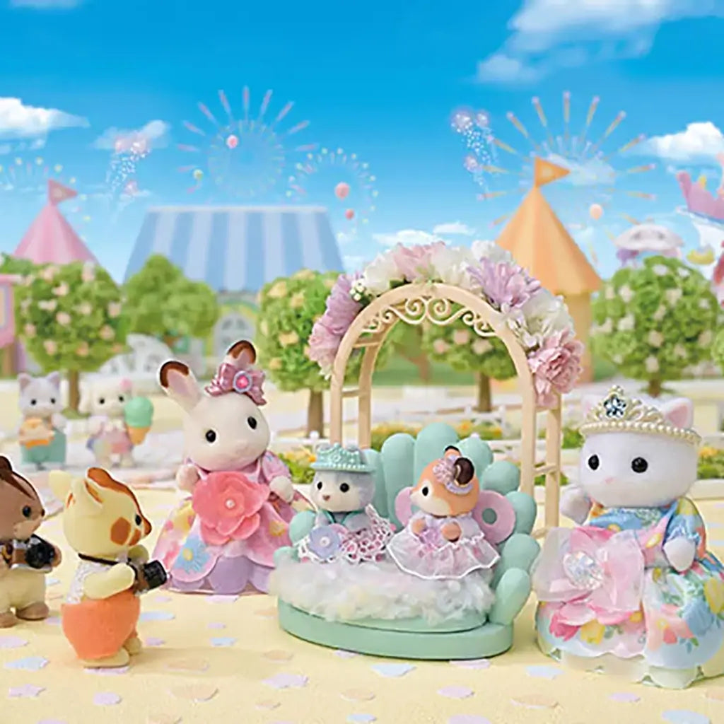 Calico Critters toy figures dressed in pastel princess gowns and tiaras are arranged on a floral stage under a sunny sky with carnival tents and trees in the background.