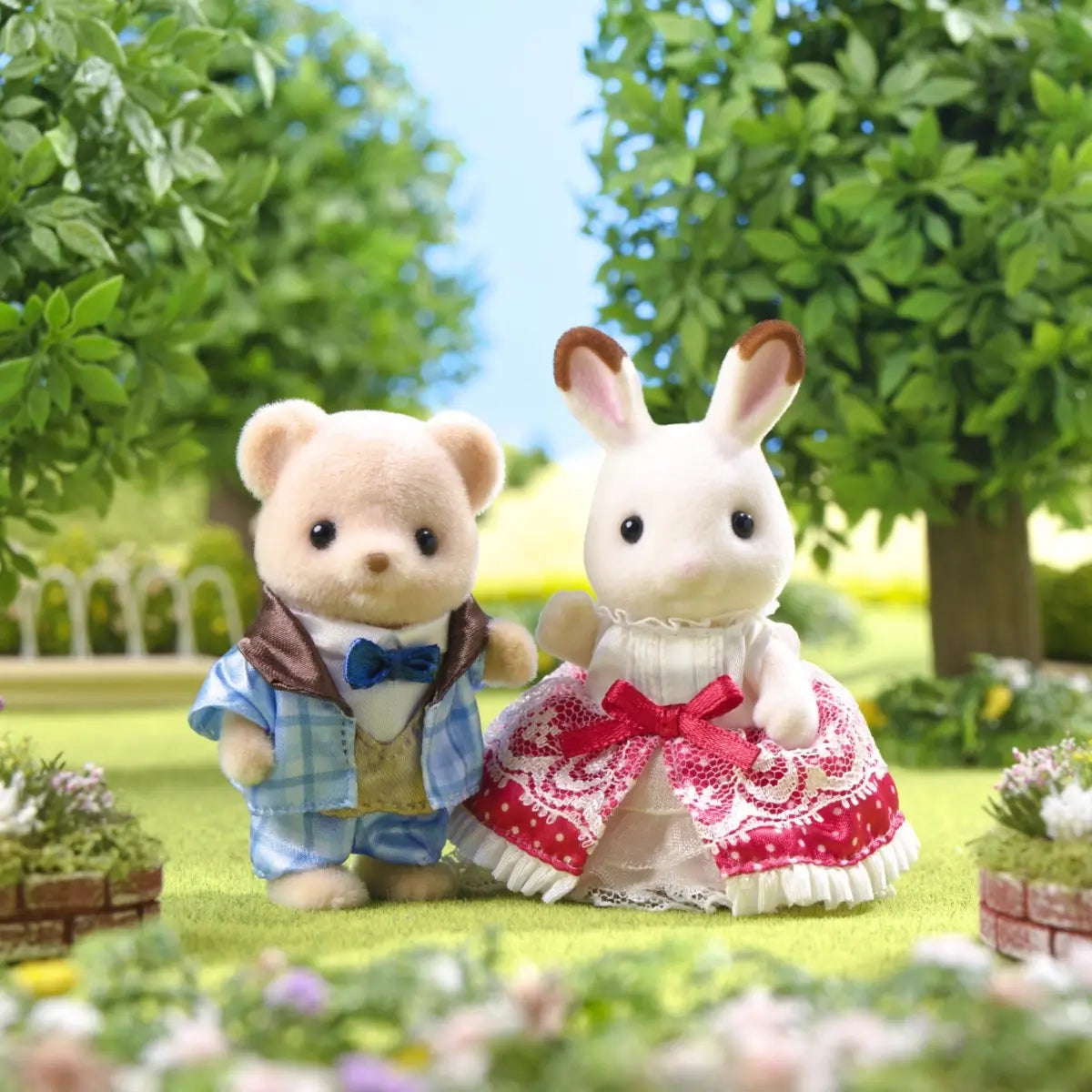Calico Critters Freya and Theo Anniversary Friends set with cream and tan flocked figures in formal outfits, lace dress with red bow and blue plaid suit, posed in green garden park setting.