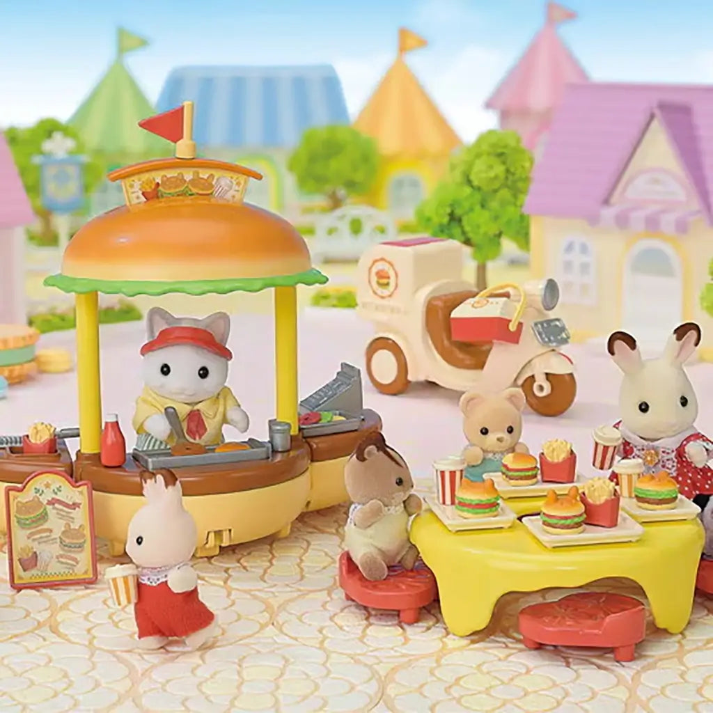 Calico Critters Hamburger Stand playset showing animal figures dining at a yellow table with burgers, fries, and drinks while a cat vendor serves food under a burger-shaped canopy.