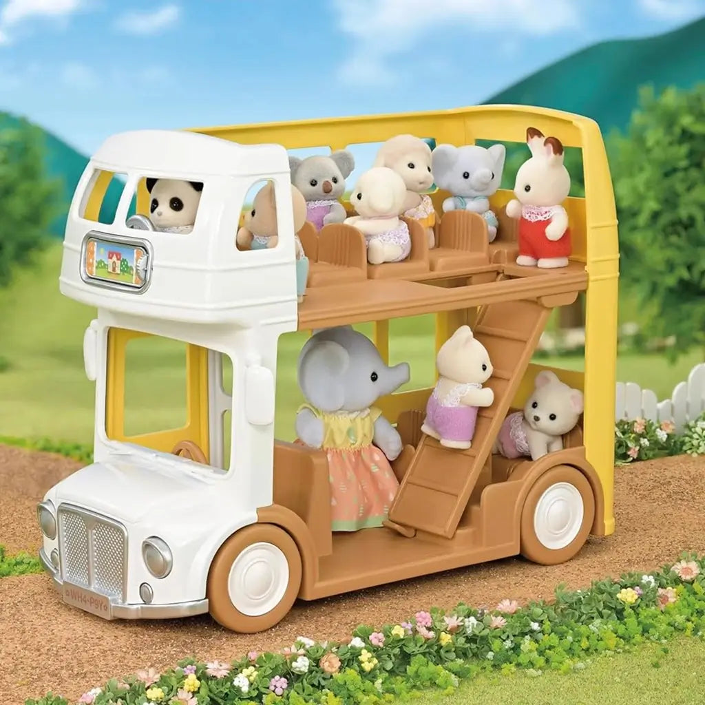Calico Critters yellow and white double-decker bus filled with small animal characters climbing stairs and sitting on brown seats, shown on a garden path surrounded by flowers and green hills.
