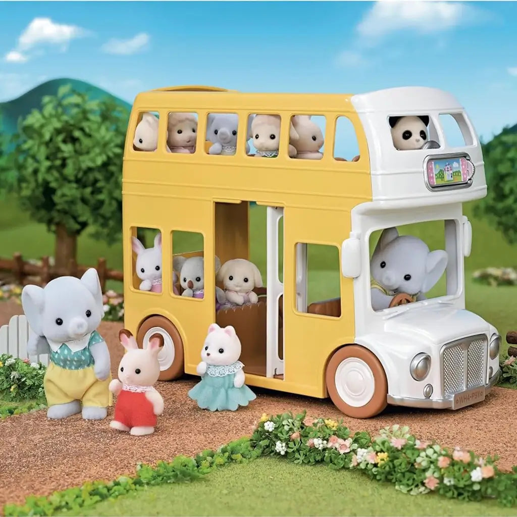 Calico Critters Nursery Double Decker Bus in yellow and white carrying multiple animal figures including rabbits, elephants, and bears, set in a grassy outdoor scene with trees and mountains in the background.