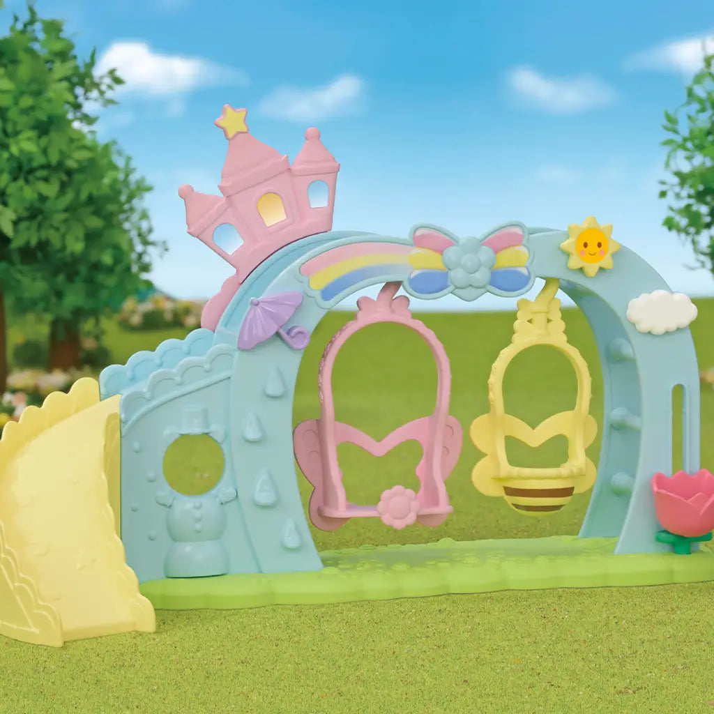 Calico Critters Nursery Swing playset outdoors showing pastel castle arch with rainbow, slide, and swings under blue sky and green grass background.
