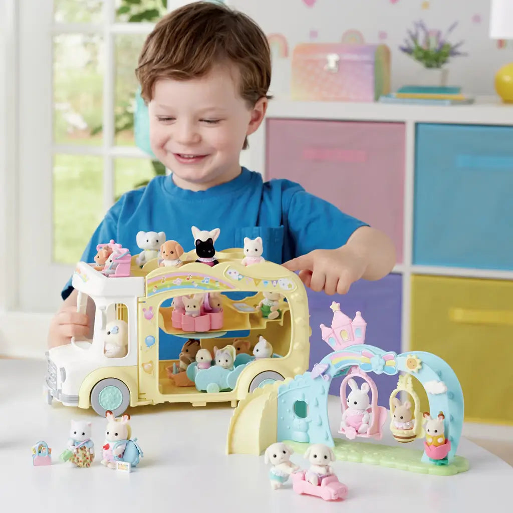 Smiling child in blue shirt plays indoors with Calico Critters Nursery Swing and nursery bus, arranging miniature animal figures on pastel play structures.