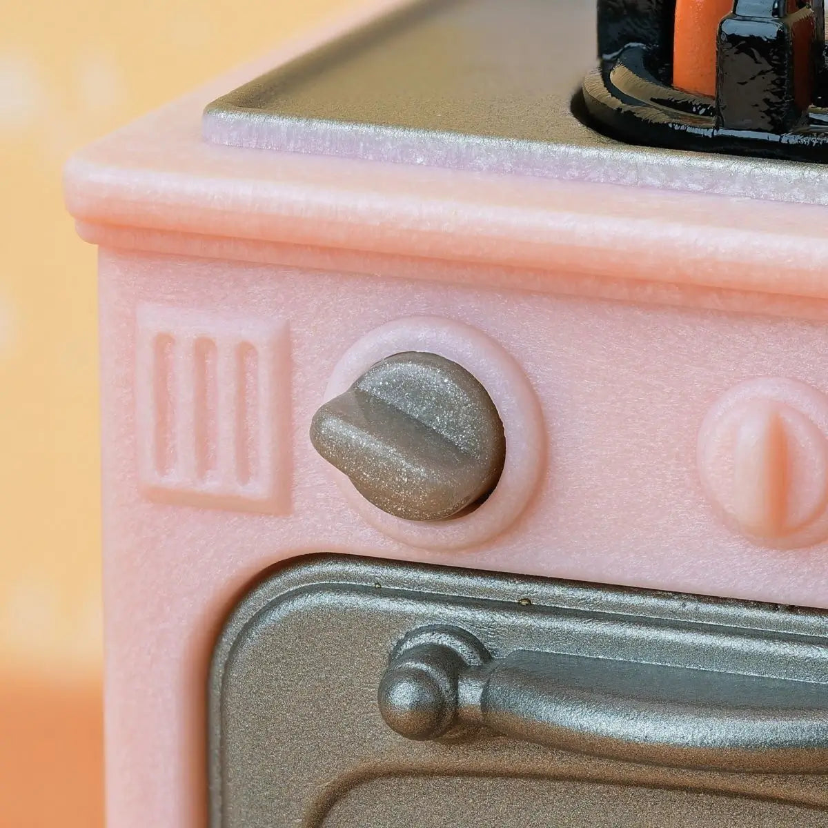 Calico Critters pink stove and oven unit features pastel pink plastic body with silver knobs, stovetop burner, and oven door with tray holding baked pie on white background.