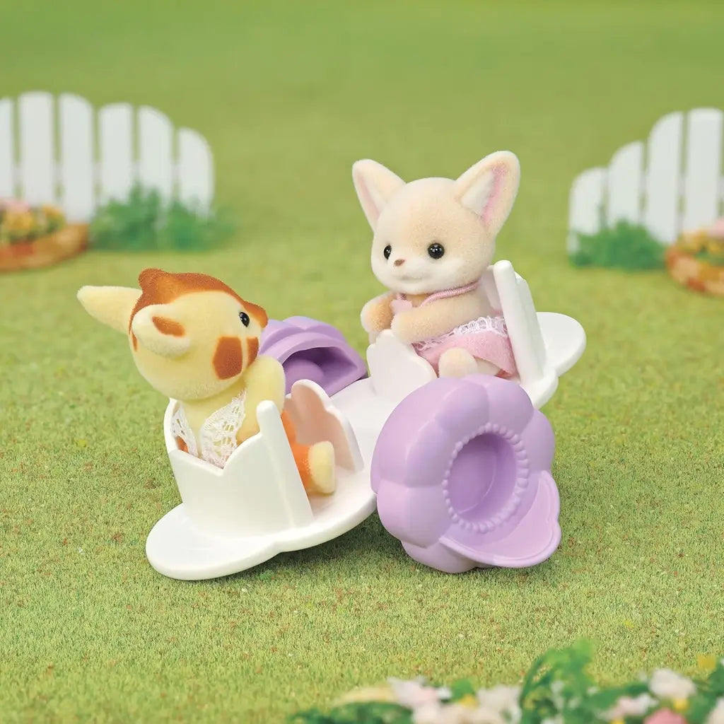 Calico Critters Playtime Castle Nursery outdoor scene with baby figures on a lavender see-saw in a grassy garden, showing playful interaction between a fennec fox and giraffe character.