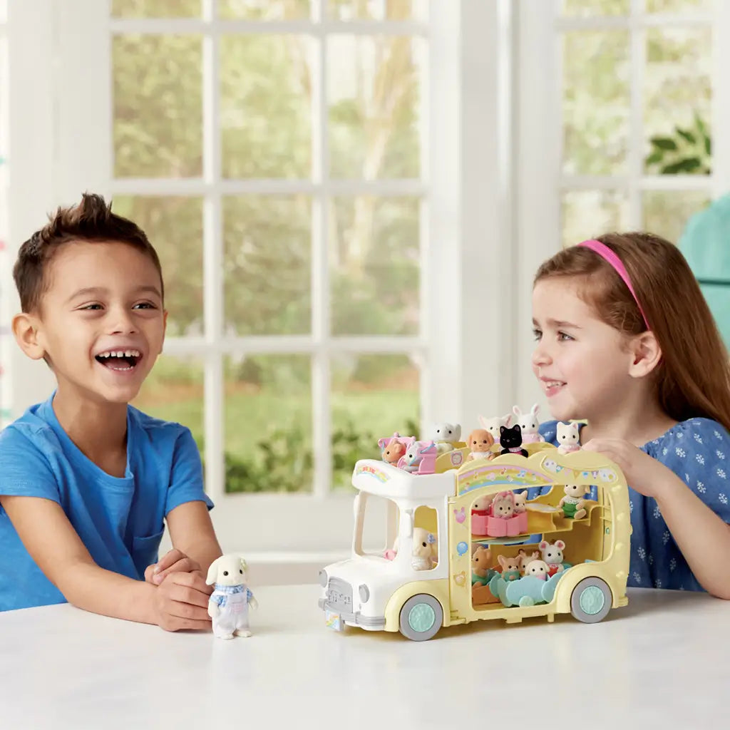 Two smiling children play indoors with the Calico Critters Rainbow Fun Nursery Bus filled with baby animal figures, pastel seats, and rotating play features.