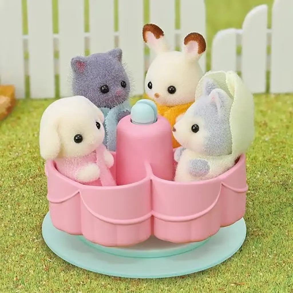Four Calico Critters baby animal figures ride on pink merry-go-round accessory included with the Rainbow Fun Nursery Bus, set outdoors on grassy ground.