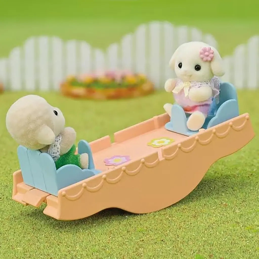Two Calico Critters baby figures sit opposite each other on orange seesaw accessory included with Rainbow Fun Nursery Bus playset in grassy yard setting.