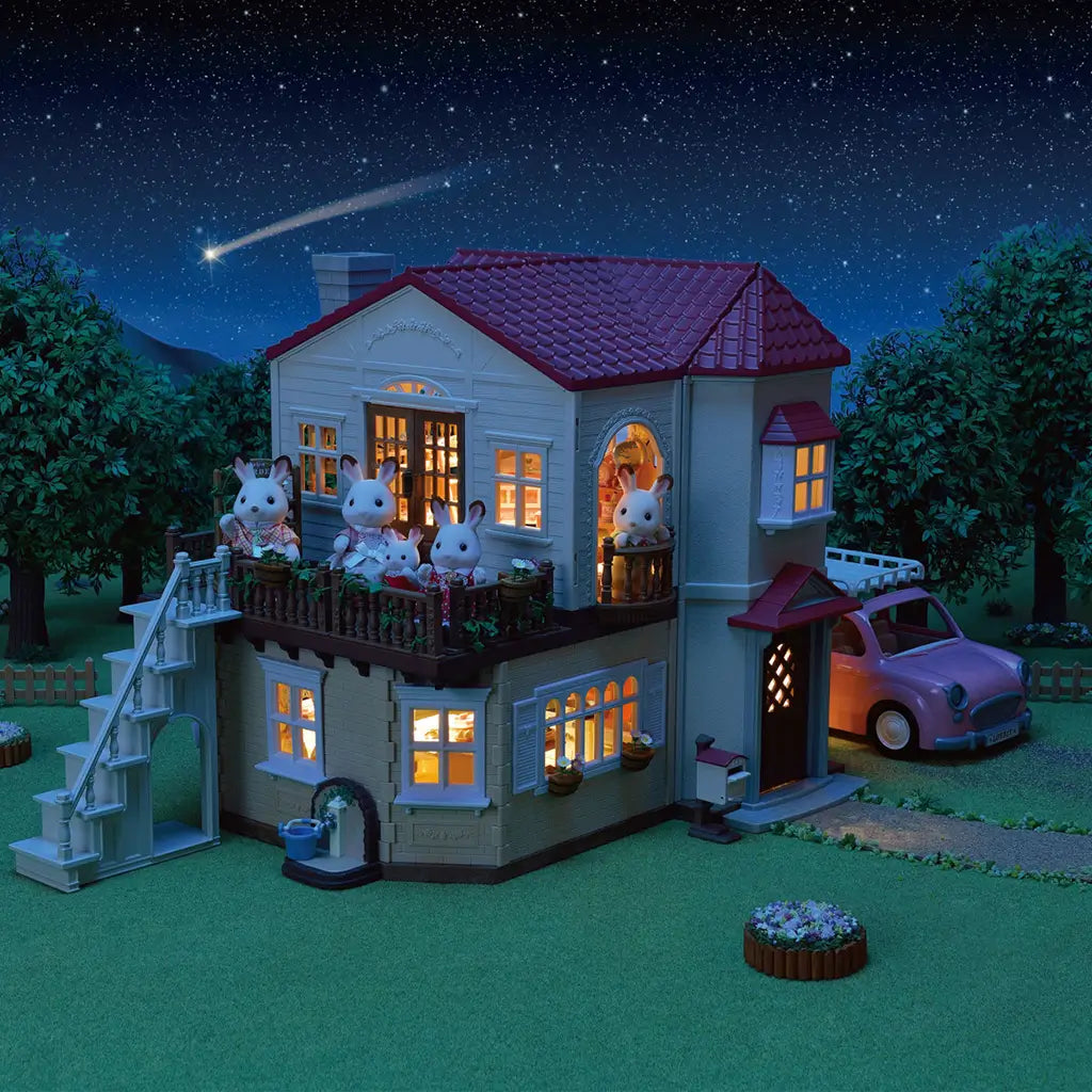 Calico Critters Red Roof Country Home shown at night with warm glowing lights, rabbit family figures on the balcony, trees, and a pink toy car in a starry outdoor setting.