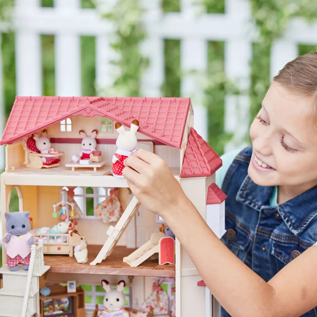 Smiling child playing with Calico Critters Red Roof Country Home, placing rabbit figure inside dollhouse with red roof, multiple rooms, and miniature toy animals and furniture.