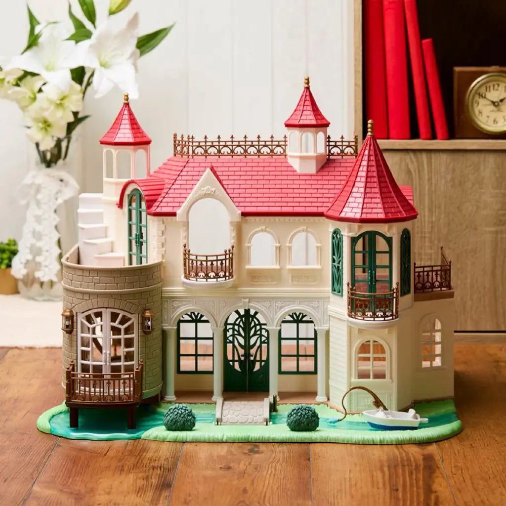 Calico Critters Red Roof Riverside Manor features white molded walls, red turret roofs, green doors, bronze balconies, river base, shrubs, and a small fishing boat.