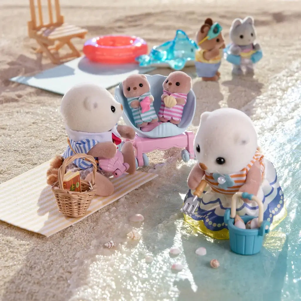 Calico Critters Sea Otter Family enjoying beach play scene with babies in stroller, father holding basket, and mother collecting shells in bucket, with other critters in background by sea.