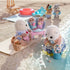 Calico Critters Sea Otter Family enjoying beach play scene with babies in stroller, father holding basket, and mother collecting shells in bucket, with other critters in background by sea.
