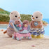 Calico Critters Sea Otter Family figures on sandy beach with stroller, two baby otters in seashell stroller, and parents dressed in striped outfits, with water and greenery in background.