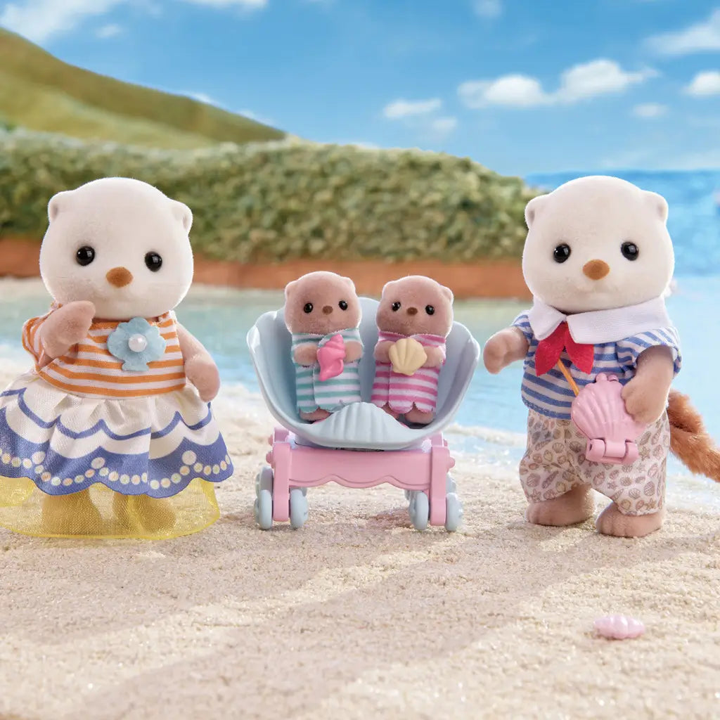 Calico Critters Sea Otter Family posed by ocean waves with twins in seashell stroller, father in striped shirt holding shell toy, and mother in striped dress, against a scenic beach backdrop.