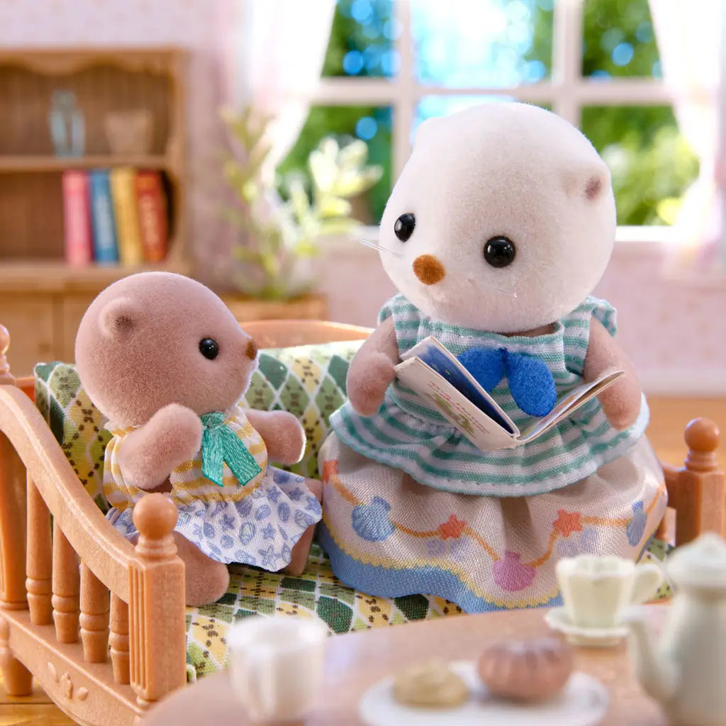 Calico Critters Sea Otter Sisters sitting on a patterned wooden sofa in a cozy living room, with the older sister reading a book to the baby under bright daylight through the window.