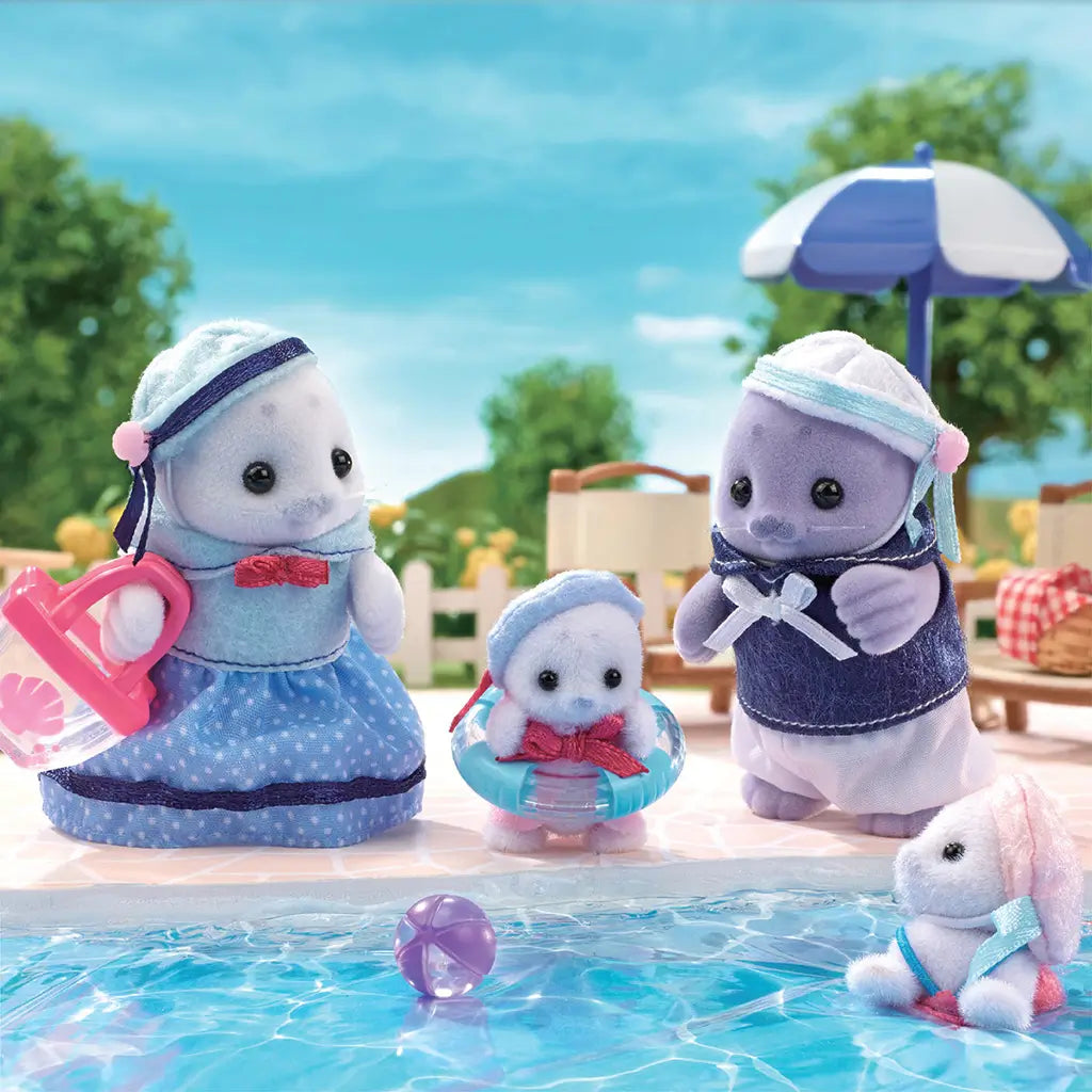 Toy Calico Critters Seal Family figurines posed at the poolside with umbrella, mother seal carrying tote, baby seals playing with float ring and ball, set against blue sky.