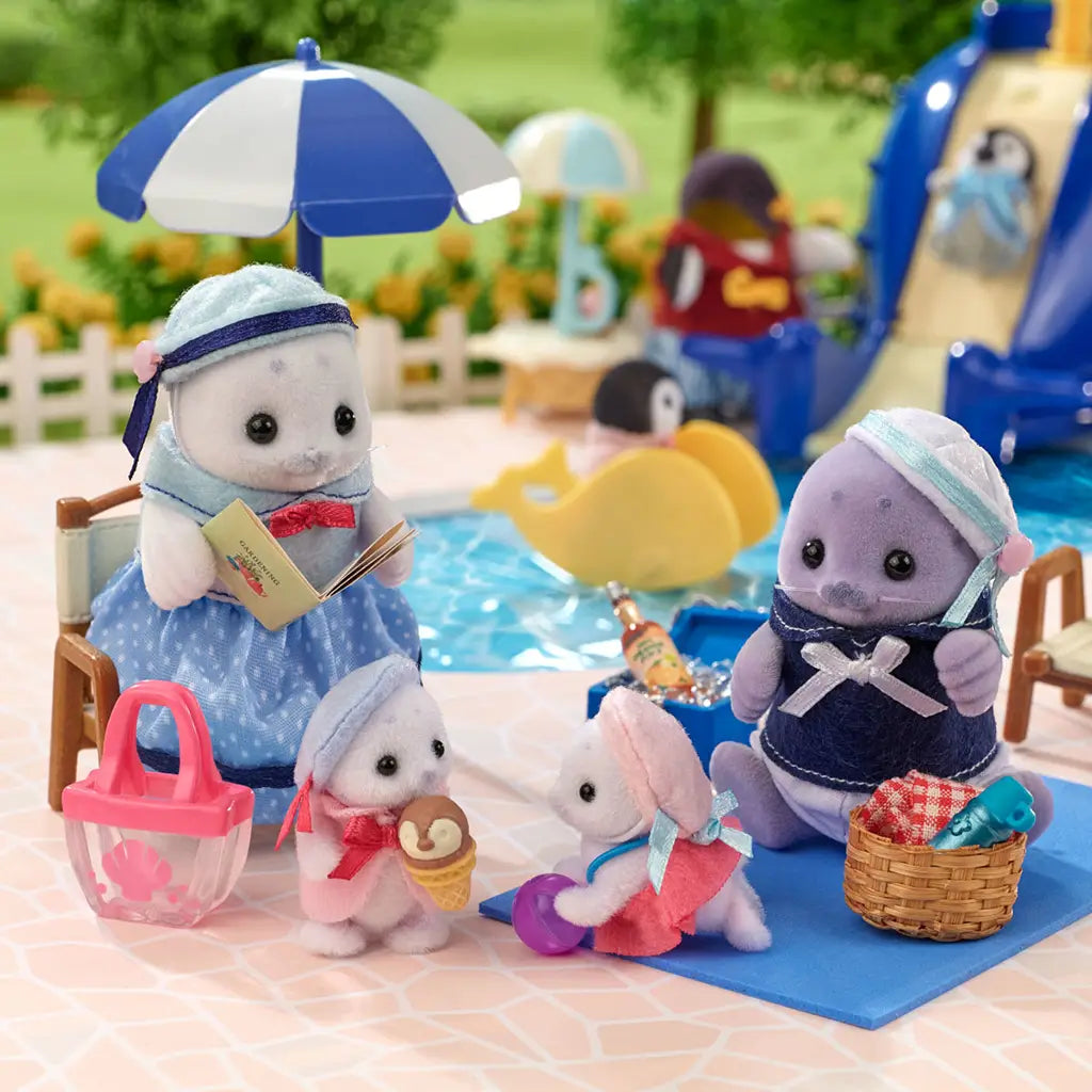 Calico Critters Seal Family figurines posed at a toy poolside with umbrella, basket, and ice cream, dressed in playful blue and pink outfits, creating a summer play scene.