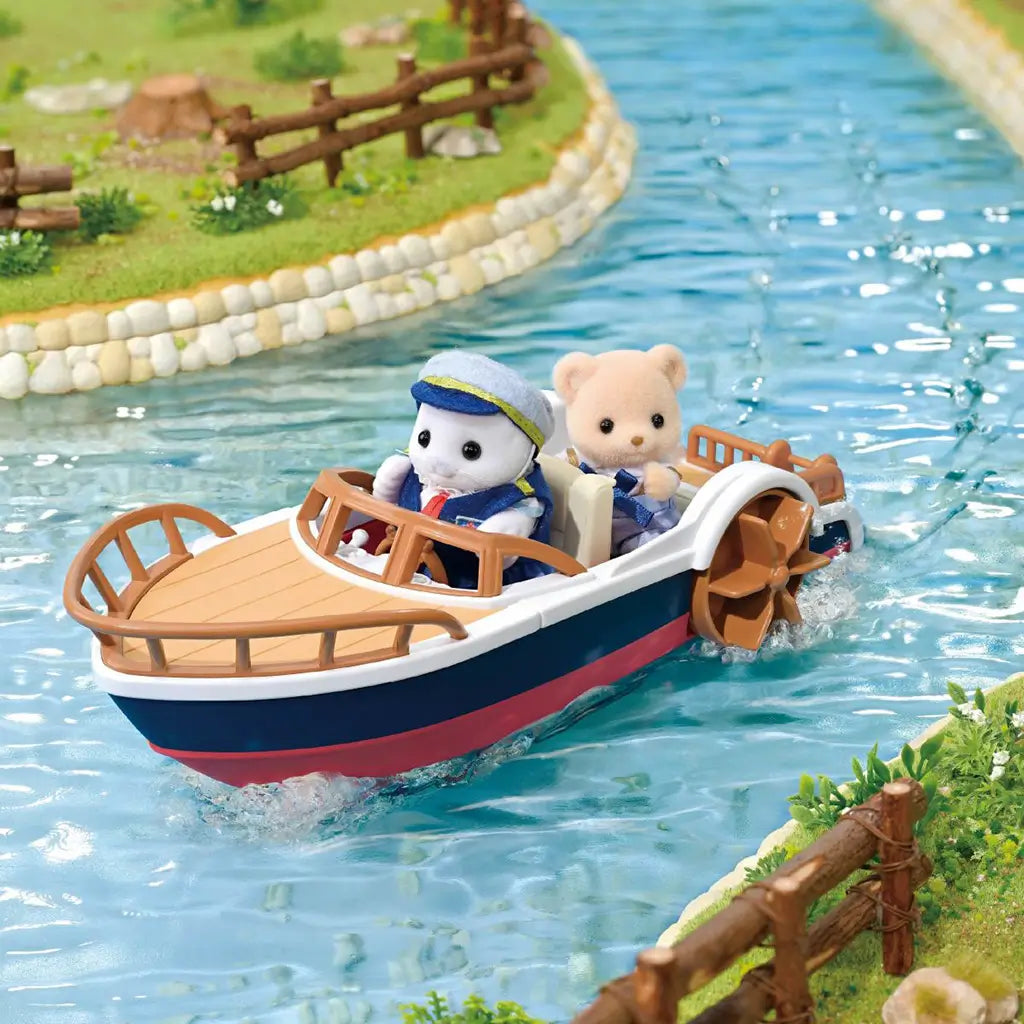 Calico Critters Seal Sailor's Leisure Boat scene shows a navy, white, and red paddle boat carrying two figures through turquoise water beside grassy, fenced riverbanks.