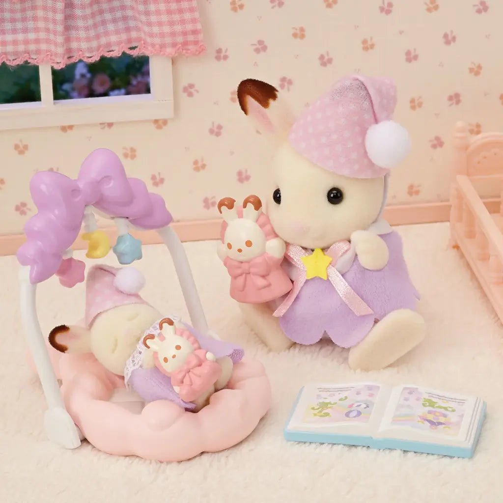 Calico Critters Sleepy Dream Siblings toy set showing a big bunny in purple pajamas holding a doll next to a baby bunny in a pink cradle, with book and nursery backdrop.