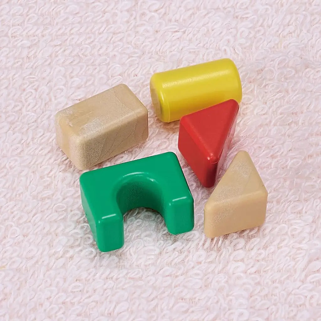 Colorful toy building blocks including red triangle, green arch, yellow cylinder, and beige shapes rest on a soft textured surface, designed for early learning and creative play.