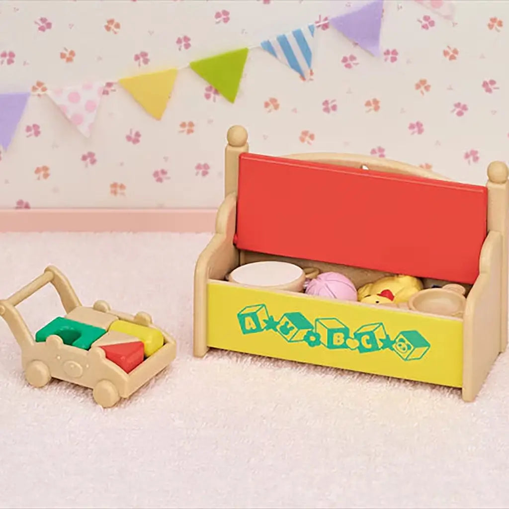 Miniature toy bench with a bright red lid and yellow front decorated with ABC blocks stands beside a small wooden cart holding colorful shapes in a nursery-themed background with bunting.