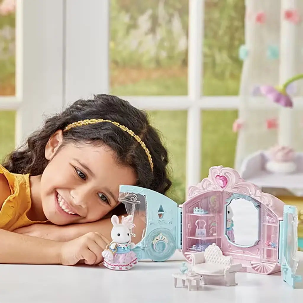 Smiling child in a yellow dress plays indoors with the Calico Critters Style & Sparkle Dressing Room set, featuring a rabbit doll, carriage-shaped case, sofa, and mirror.