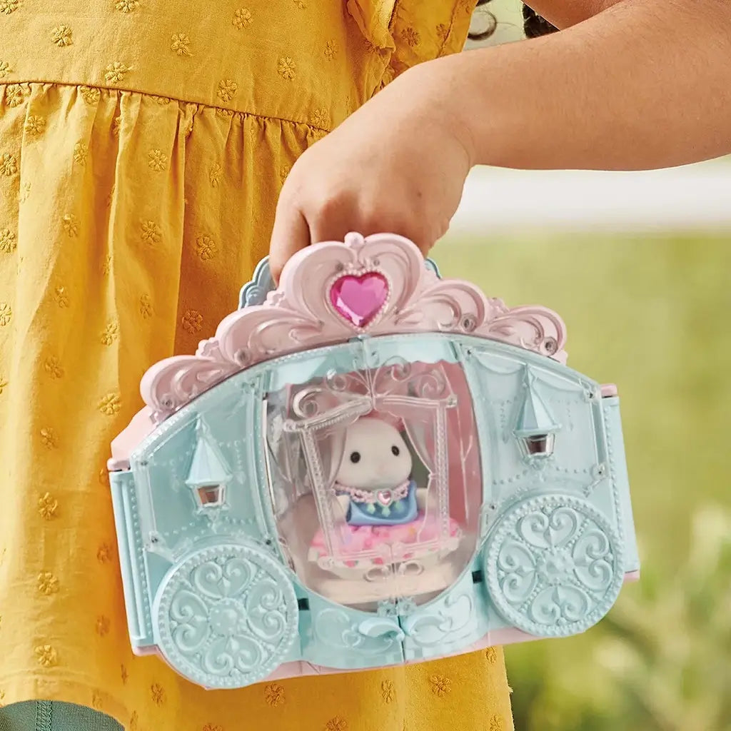 Toy playset carriage in pastel blue and pink with ornate heart details, featuring a Calico Critters rabbit doll inside, held by a child wearing a mustard yellow dress outdoors.