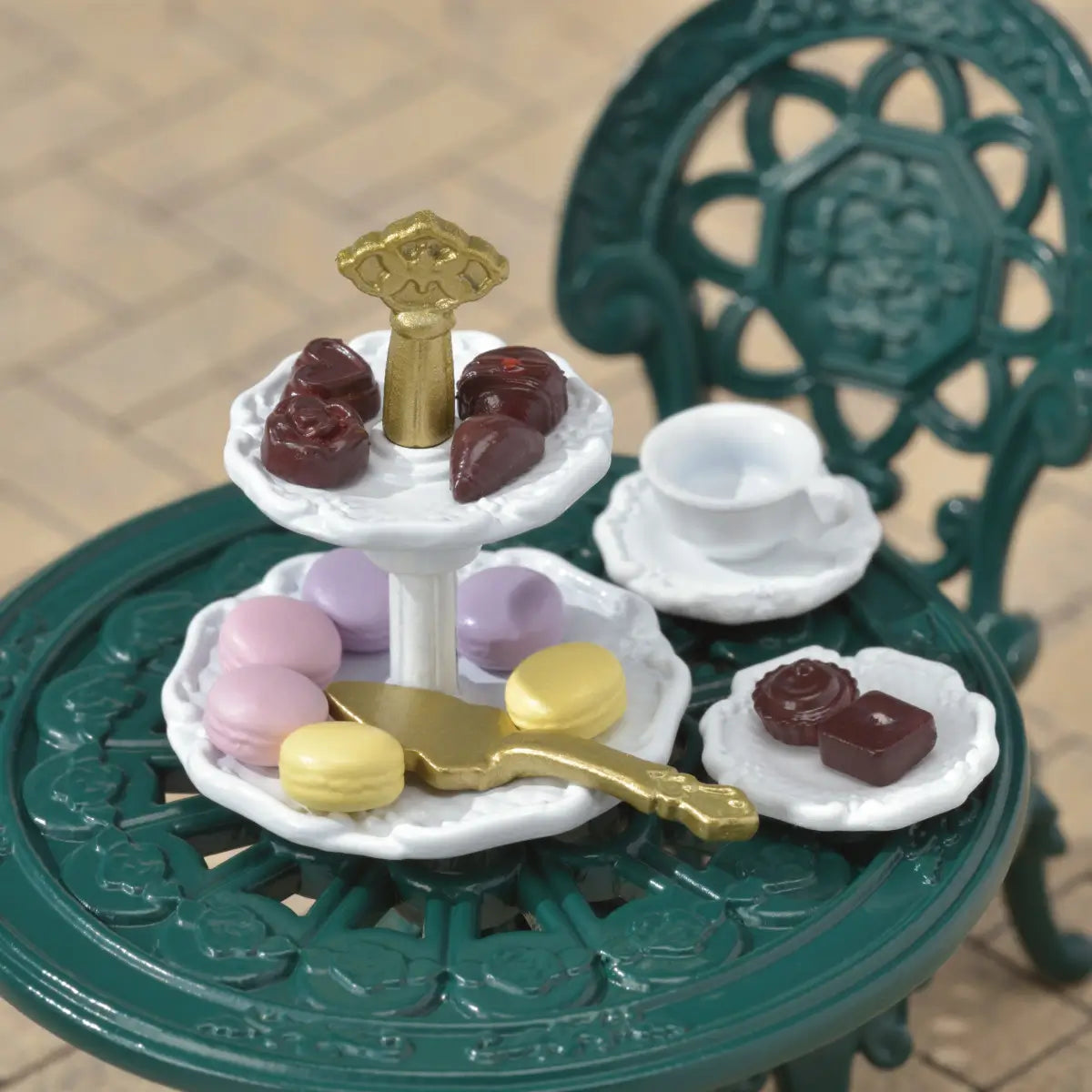 Calico Critters Tea and Treats set features dark green ornate plastic table and chairs with white tiered dessert stand, pastel macarons, chocolate candies, teacup, and gold server on indoor floor background.