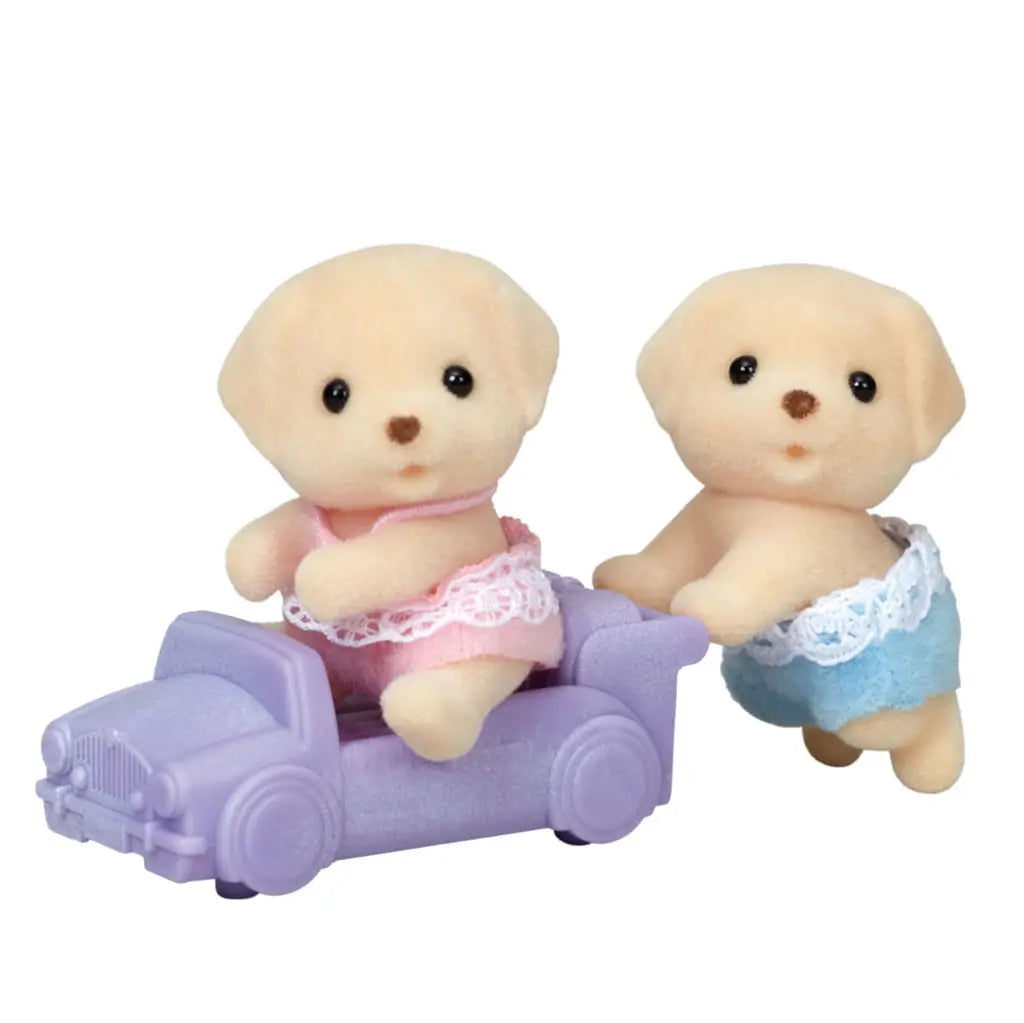 Calico Critters Yellow Labrador twin figurines with soft beige fur, one in a pink lace outfit sitting in a lavender toy car and the other in blue lace outfit pushing it.