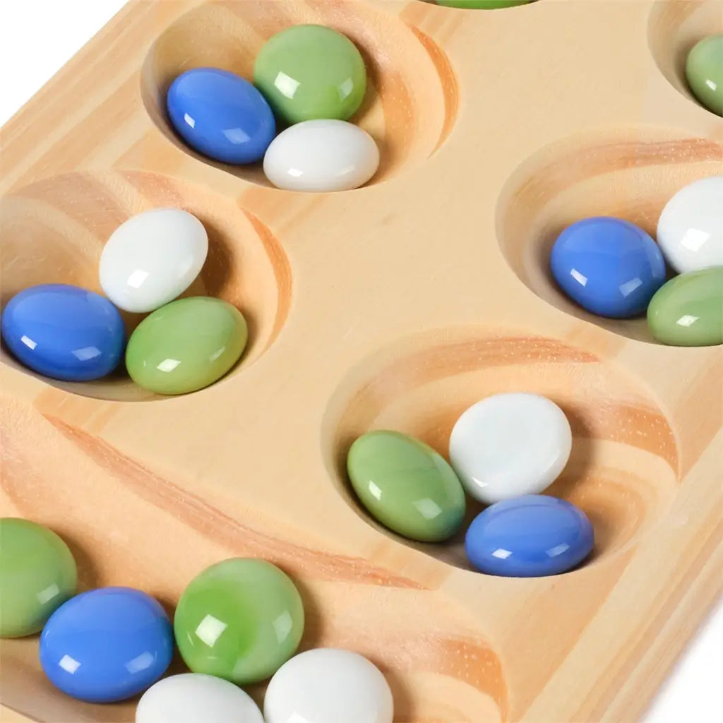 Close-up of a wooden Mancala board with smooth carved pits holding glossy blue, green, and white glass stones, highlighting the tactile texture and natural wood grain detail.