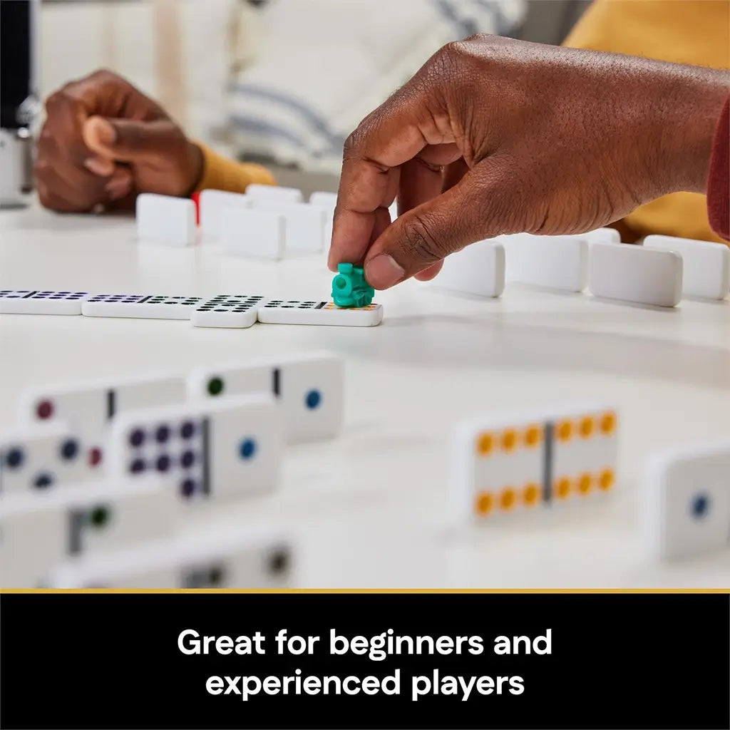 Cardinal Classics Mexican Train Dominoes gameplay with white tiles arranged in lines as a player places a green train marker by hand on a tabletop, highlighting interactive family board game action indoors.