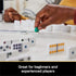 Cardinal Classics Mexican Train Dominoes gameplay with white tiles arranged in lines as a player places a green train marker by hand on a tabletop, highlighting interactive family board game action indoors.