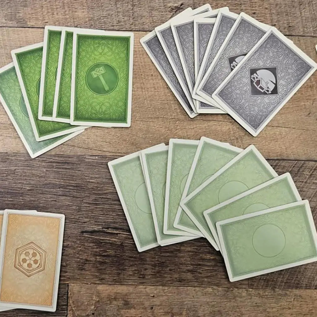 CATAN On the Road card game cards feature green, gray, tan, and pale green patterned paper backs arranged in four stacks across a rustic brown wooden tabletop.
