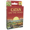 CATAN On the Road card game features red cardboard packaging with yellow title lettering, a stylized island sunset landscape, age 10+ icons, and Asmodee branding.