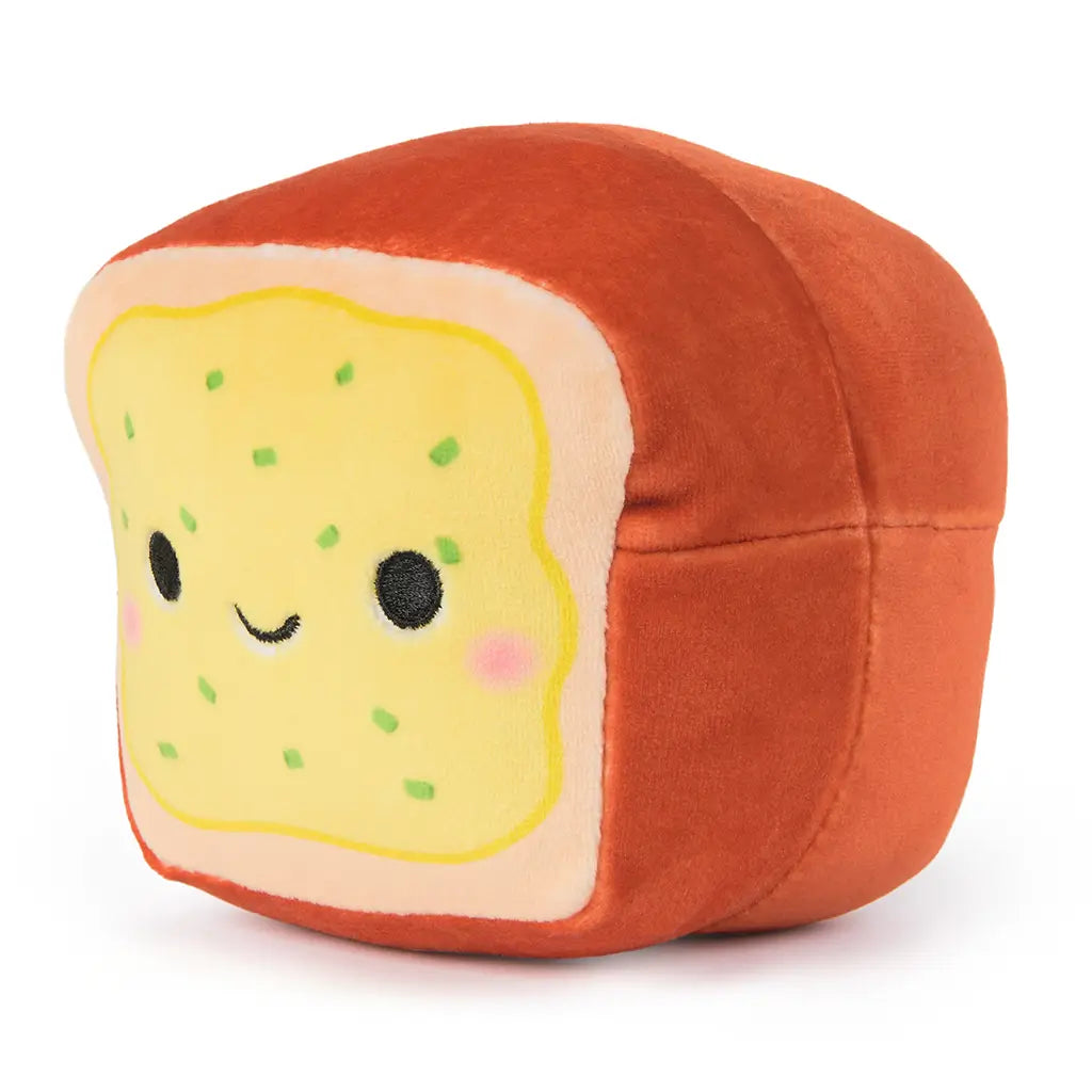 Angled view of Cheeky Chonks Rearly Garlicky reversible plush, shaped like a slice of garlic bread with smiling face and green sprinkle details on soft yellow surface.
