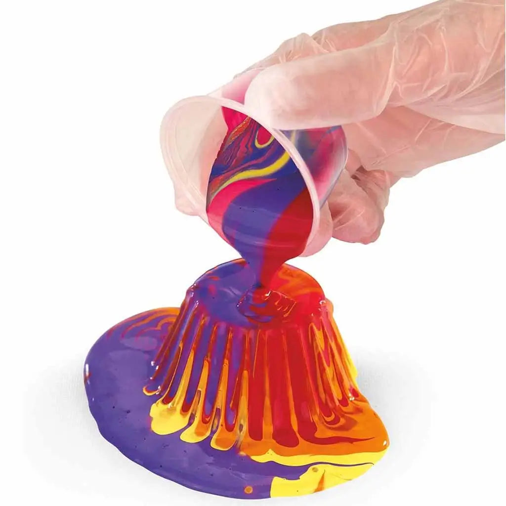 Gloved hands pouring mixed purple, red, orange, and yellow paint from a cup onto a surface, forming a layered, flowing acrylic pour pattern typical of fluid art techniques.
