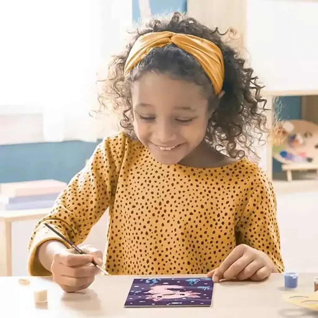 Smiling young girl painting a CreArt axolotl picture at a table, using a brush and pastel paints in a cozy, sunlit room.