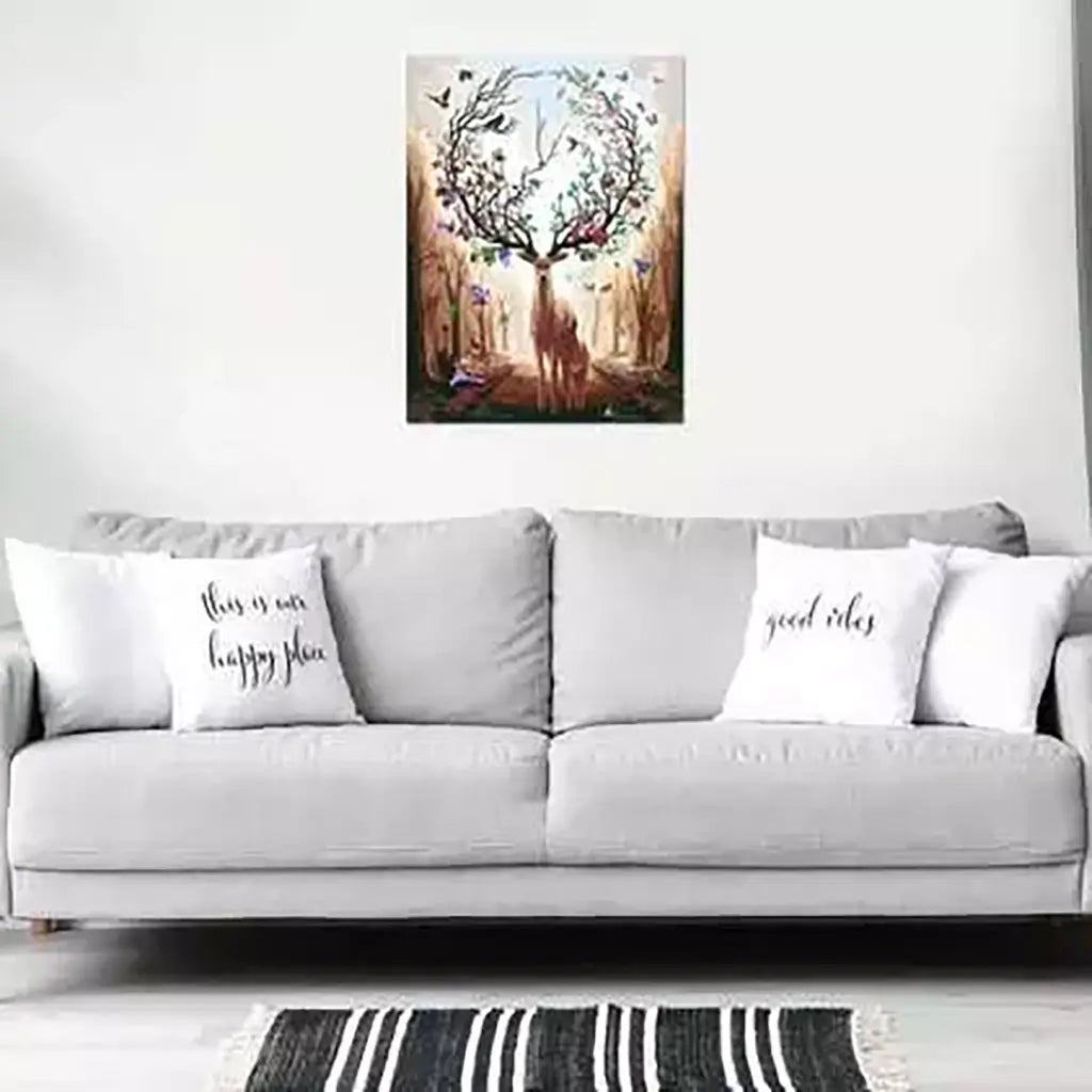 Framed paint by numbers artwork of a deer with blossoming antlers surrounded by butterflies, displayed on a white wall above a gray sofa with text pillows in a modern living room.