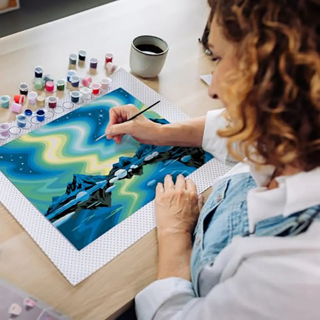 Woman painting a CreArt northern lights canvas at a table with multiple paint pots and a brush, carefully adding detail to the mountainous landscape scene.