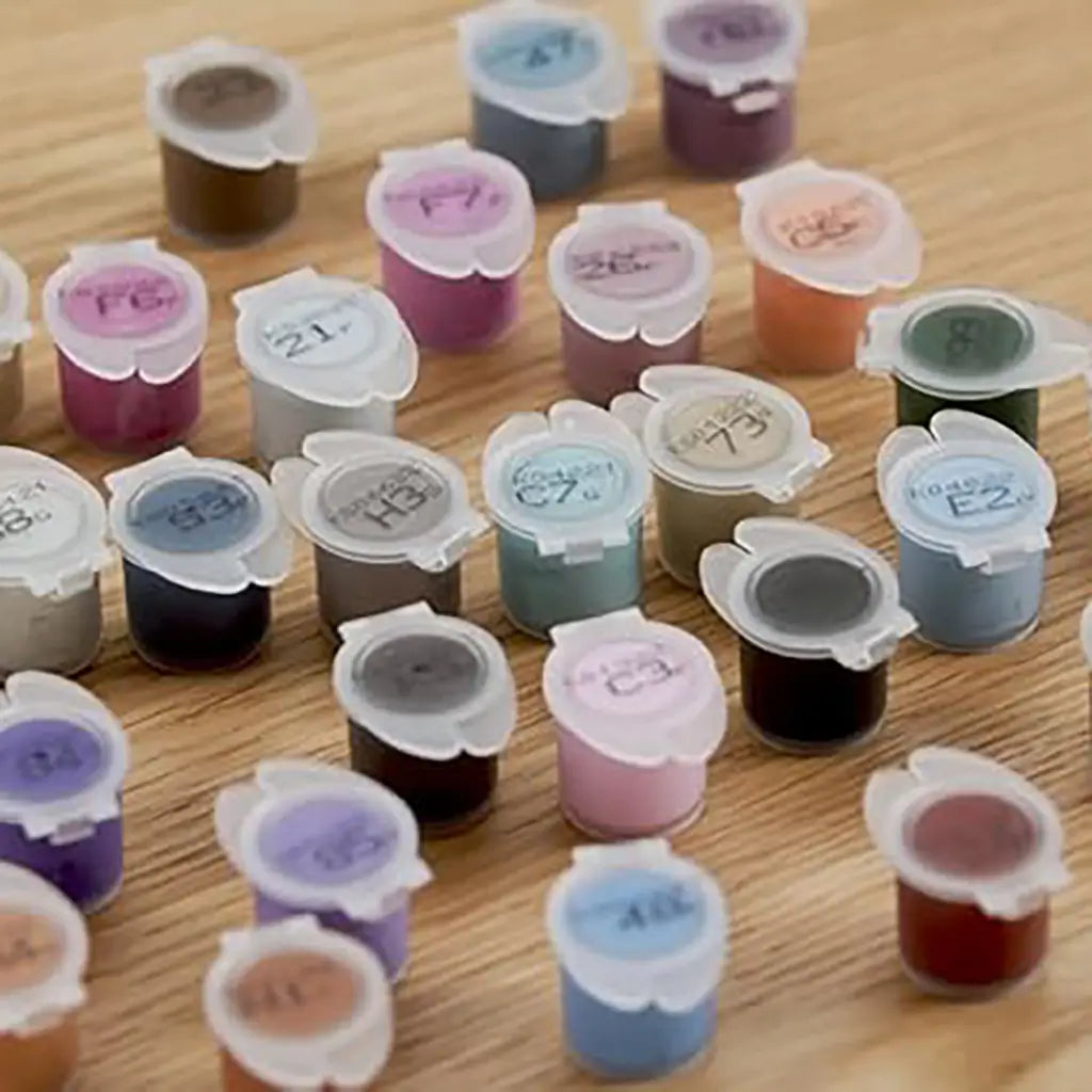 Close-up of small plastic paint pots in multiple shades like pink, green, blue, and brown, labeled with numbers for matching in the Ravensburger CreArt castle painting kit.