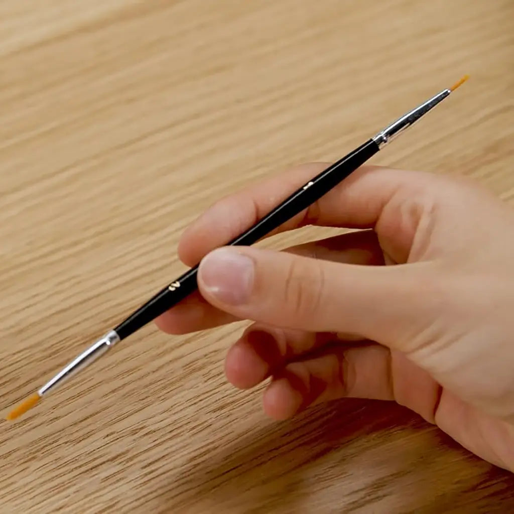 Hand holding a fine black paintbrush with a narrow orange bristle tip, included in the CreArt fairytale castle paint by numbers set for precise painting details.