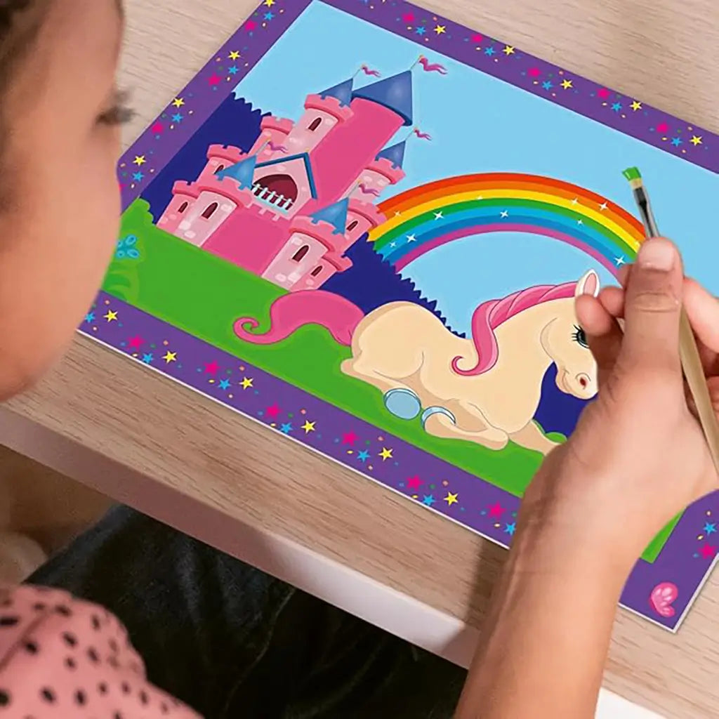 Painting by numbers art project shows a child coloring a pastel unicorn with a pink mane, rainbow, and castle scene bordered by purple frame decorated with stars.