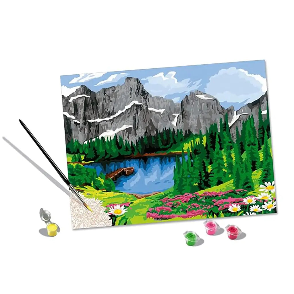 Paint by numbers canvas with brush and paints, depicting a mountain scene of tall cliffs, pine trees, clear lake, and colorful flowers being filled in.