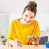 Smiling woman in a yellow shirt painting at a table with Ravensburger CreArt supplies, holding brushes and paints for a mountain landscape artwork.