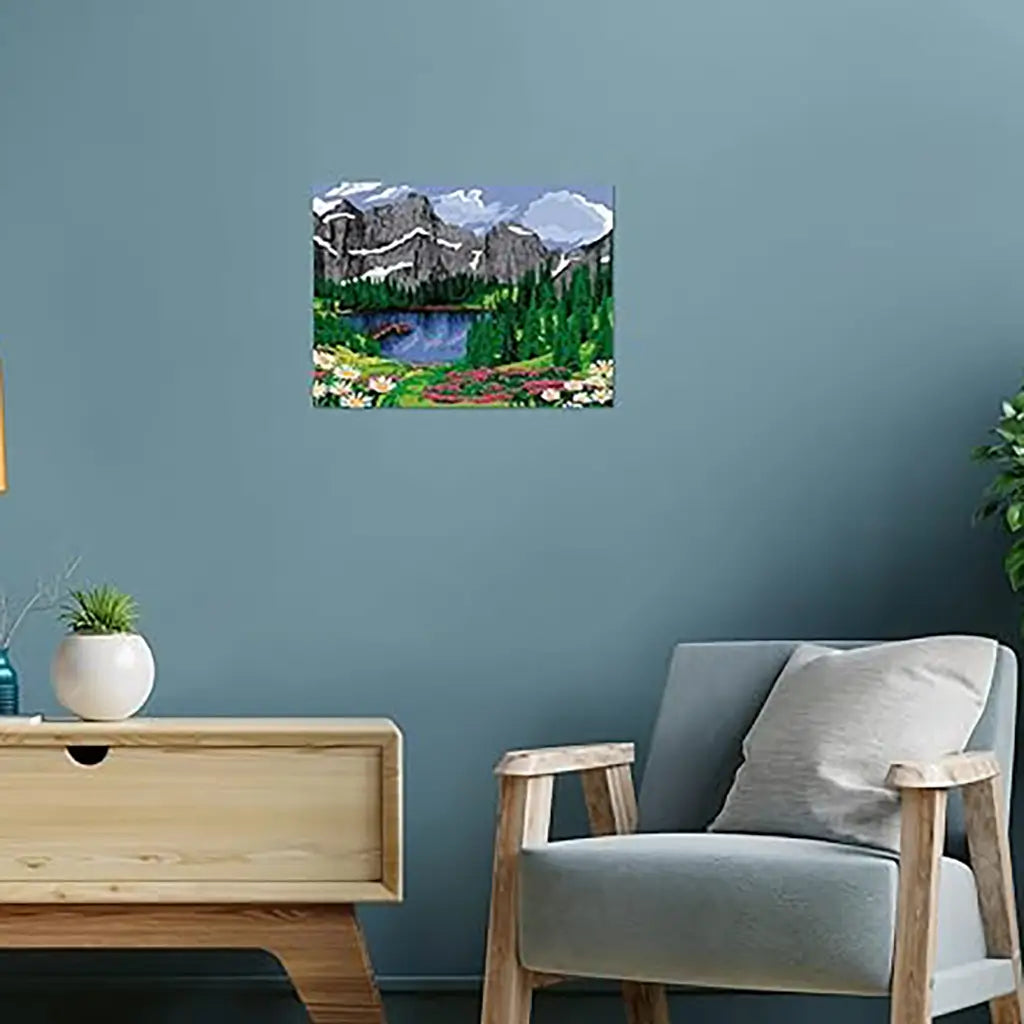 Wall art kit displayed in a modern living room, showing a vibrant mountain view painting with snowy peaks, pine trees, and a bright blue lake surrounded by flowers.