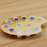 Acrylic paint palette holder with small round color pots labeled with numbers, arranged on a yellow oval board placed on a light wooden surface for organized painting use.