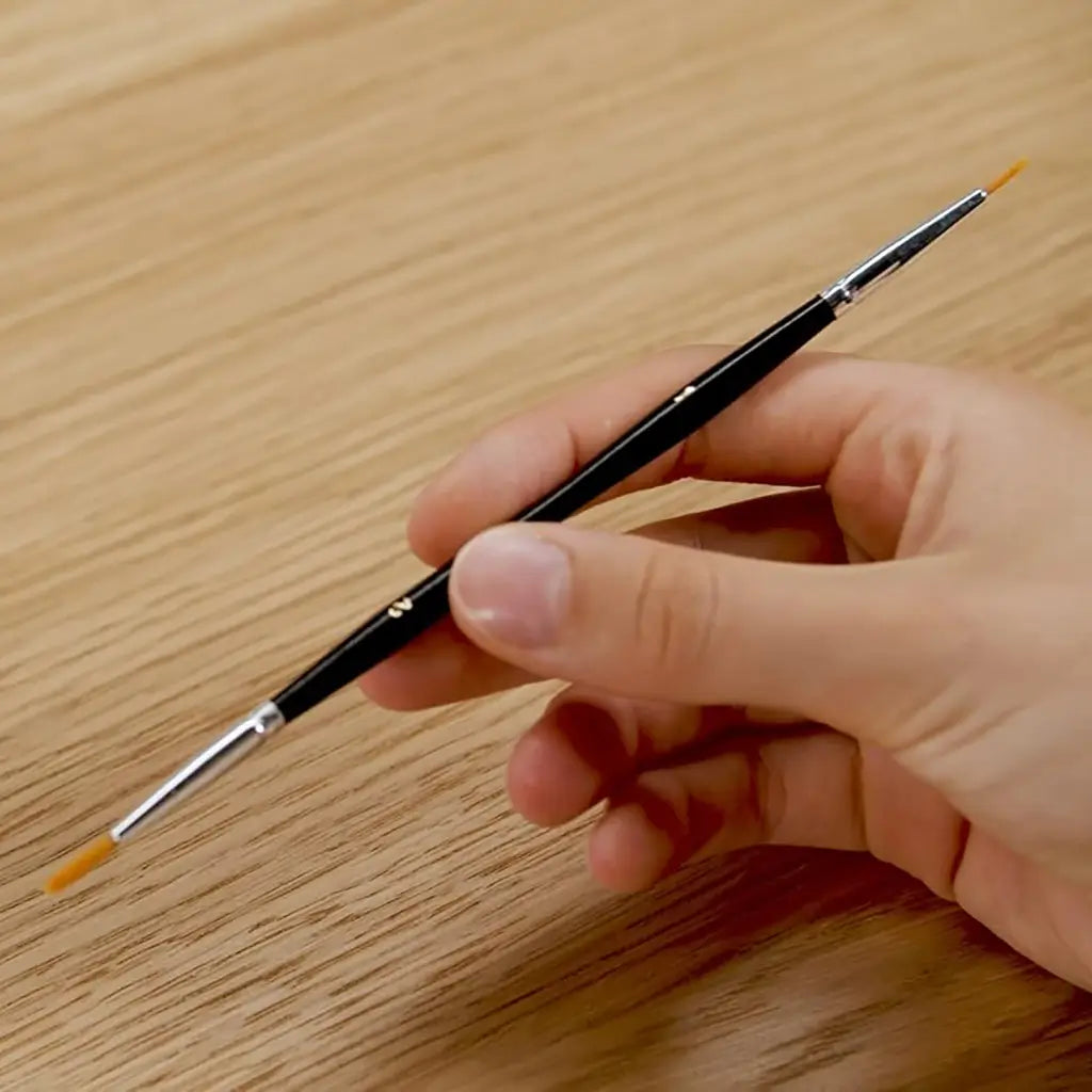 Close-up of a hand holding a slim black double-ended paintbrush with fine orange bristles, designed for detailed strokes, against a smooth light wooden table background.
