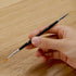 Close-up of a hand holding a slim black double-ended paintbrush with fine orange bristles, designed for detailed strokes, against a smooth light wooden table background.