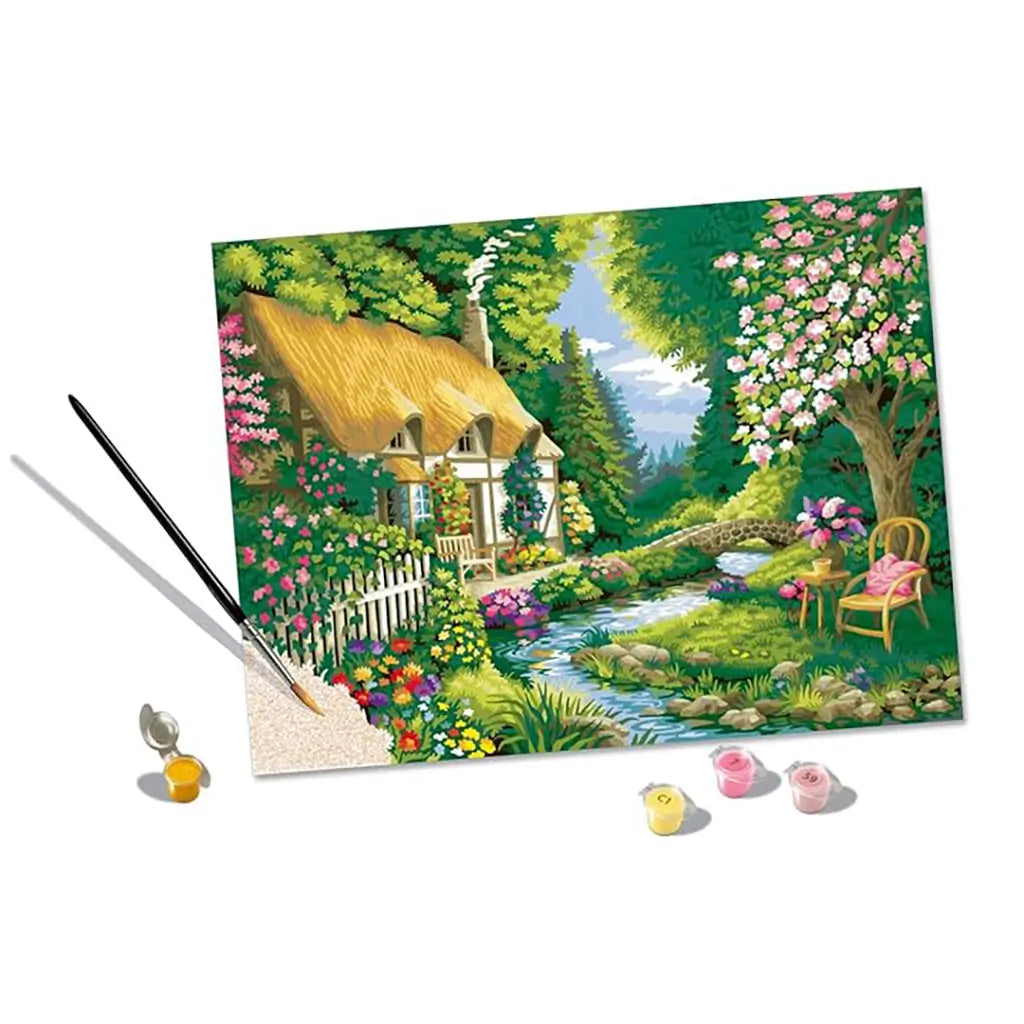 Partially completed River Cottage paint by numbers artwork with brush applying paint, showing colorful garden, stream, and trees, alongside small open acrylic paint pots.